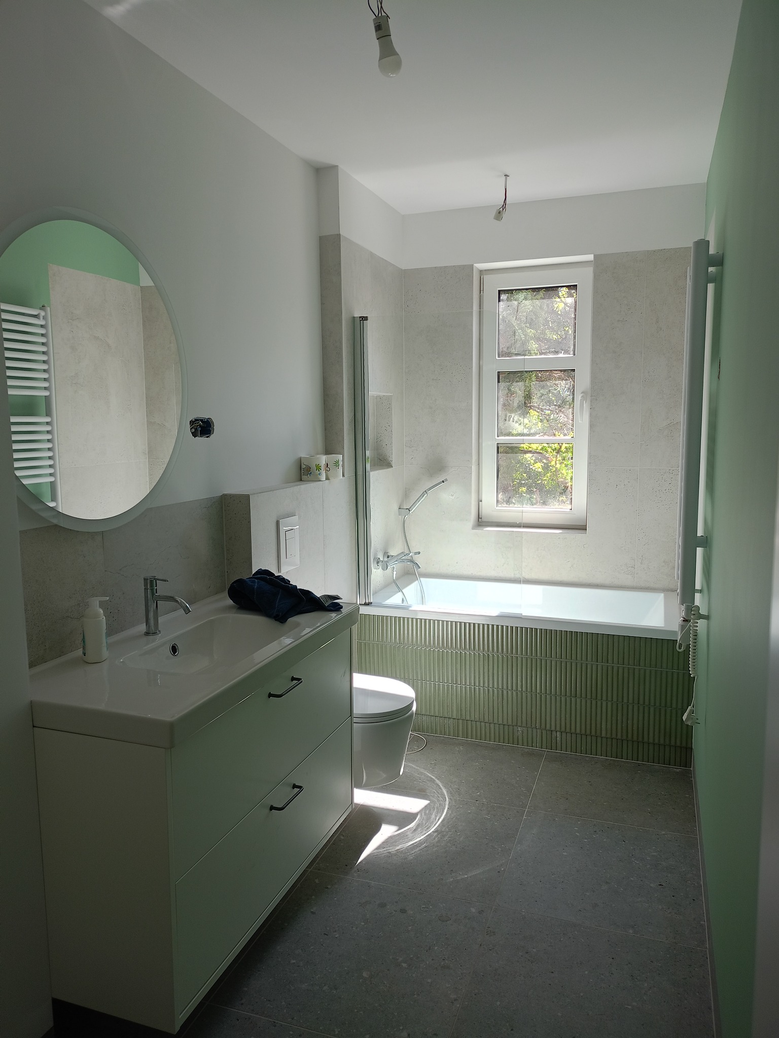 Jasna łazienka z wanną obudowaną zieloną mozaiką, okrągłe lustro nad umywalką z szafką. Minimalistyczny design, szare płytki podłogowe i naturalne światło z okna.