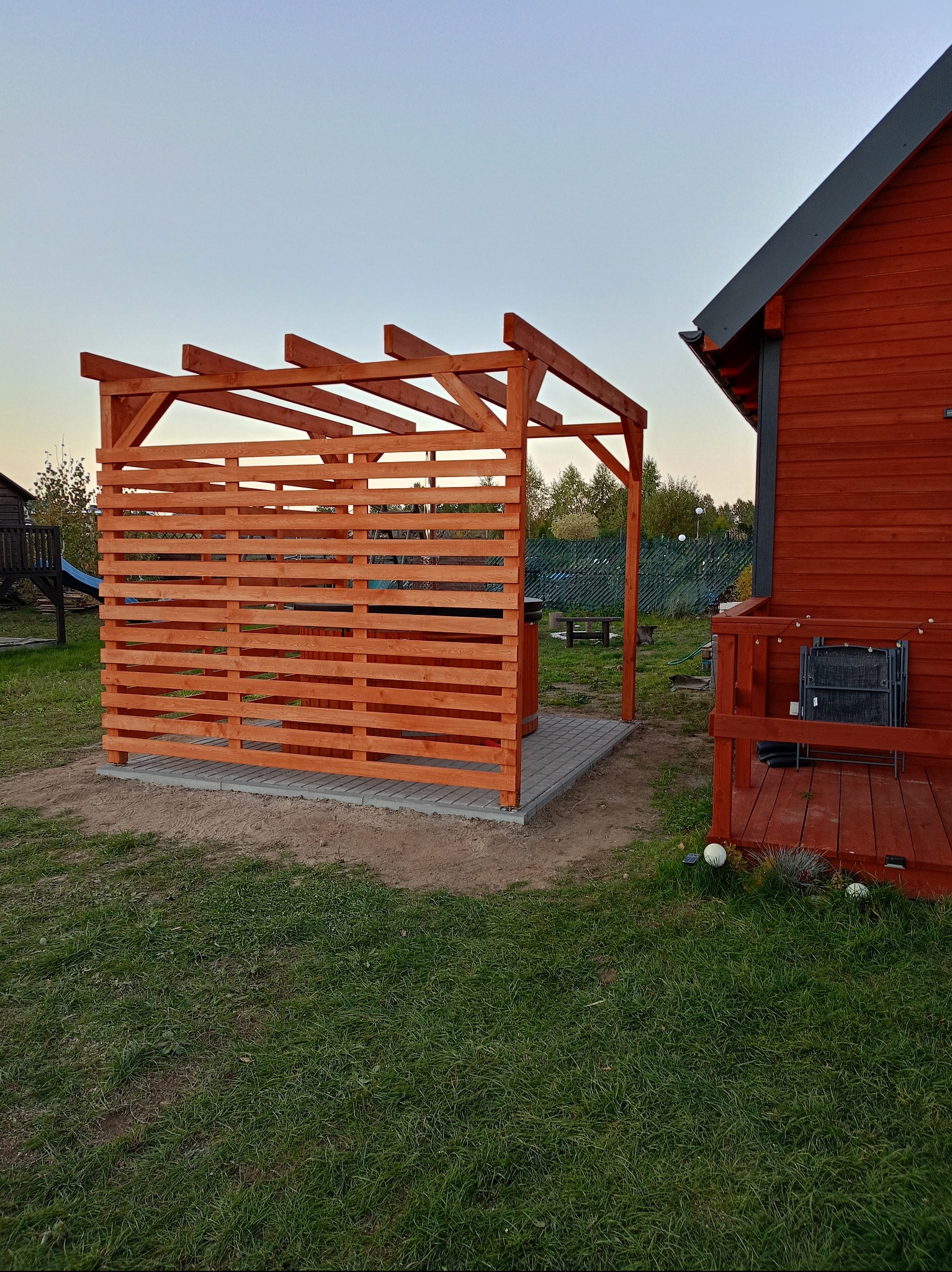Drewniana pergola z ażurową ścianą, przylegająca do domu z czerwonego drewna. Widok na ogród i elementy małej architektury w tle, w spokojnym, naturalnym otoczeniu.