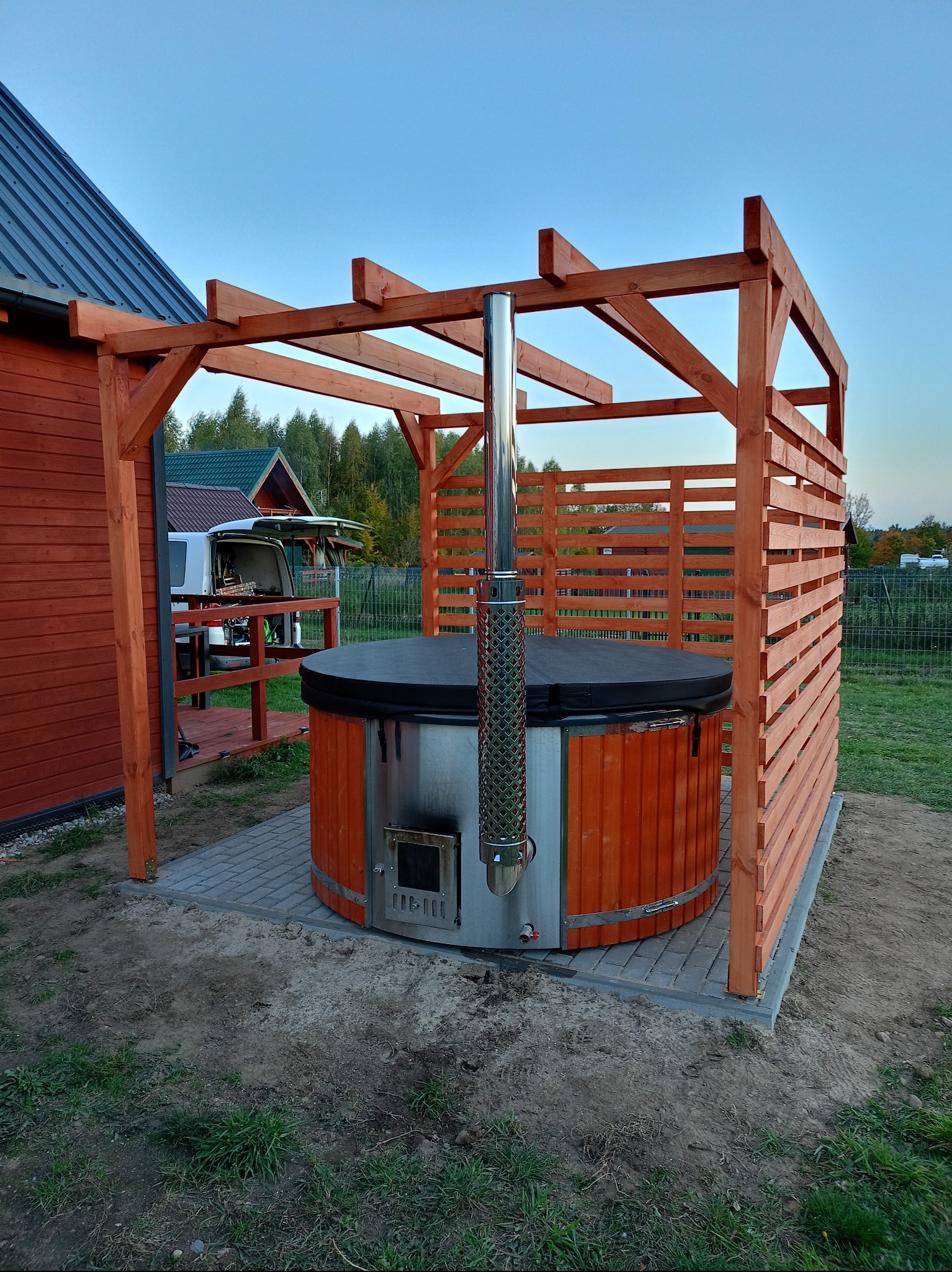 Drewniana balia z piecem na zewnątrz, otoczona drewnianą konstrukcją z płotem, na tle domu i zieleni. Widoczne detale pieca i komina.