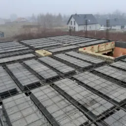 Zbrojony strop Teriva w szarej tonacji, z widocznymi pustakami betonowymi i stalową siatką. W tle zarys domów w lekkiej mgle.