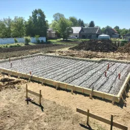 Zbrojenie fundamentów pod dom jednorodzinny. Widoczne szalunki, izolacja i wystające rury. W tle zabudowa jednorodzinna i drzewa w słoneczny dzień.