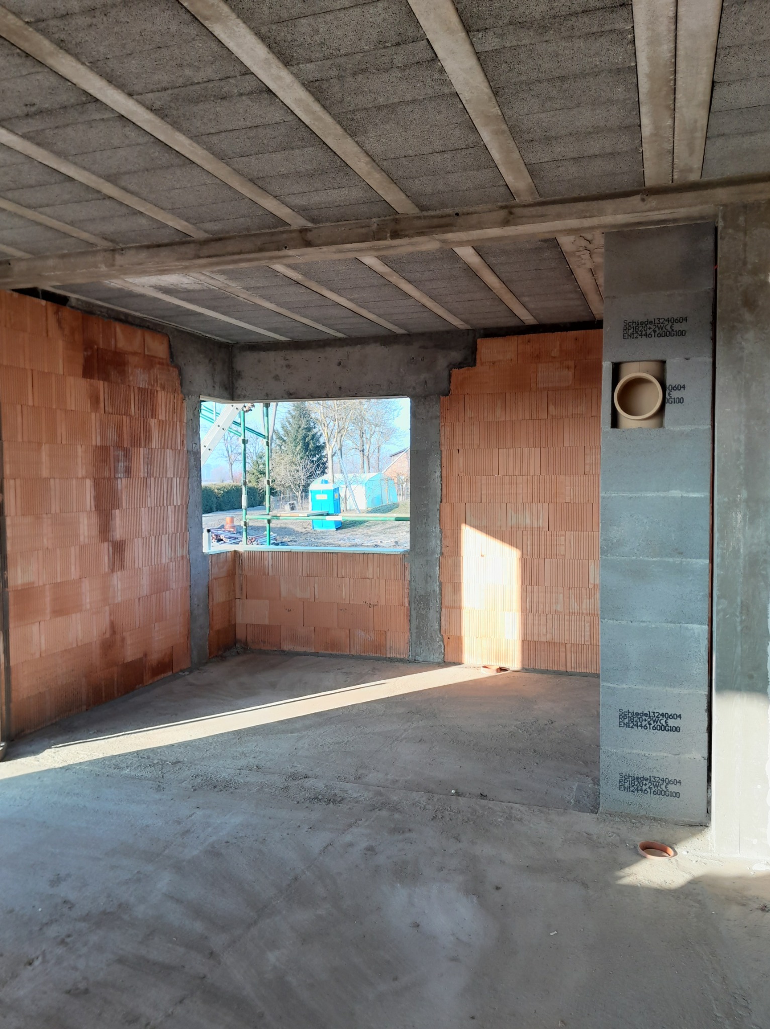 Wnętrze budynku w trakcie budowy: ściany z czerwonej cegły, surowy betonowy sufit z belkami, widoczne okno z widokiem na zewnątrz, betonowa podłoga i pion wentylacyjny.