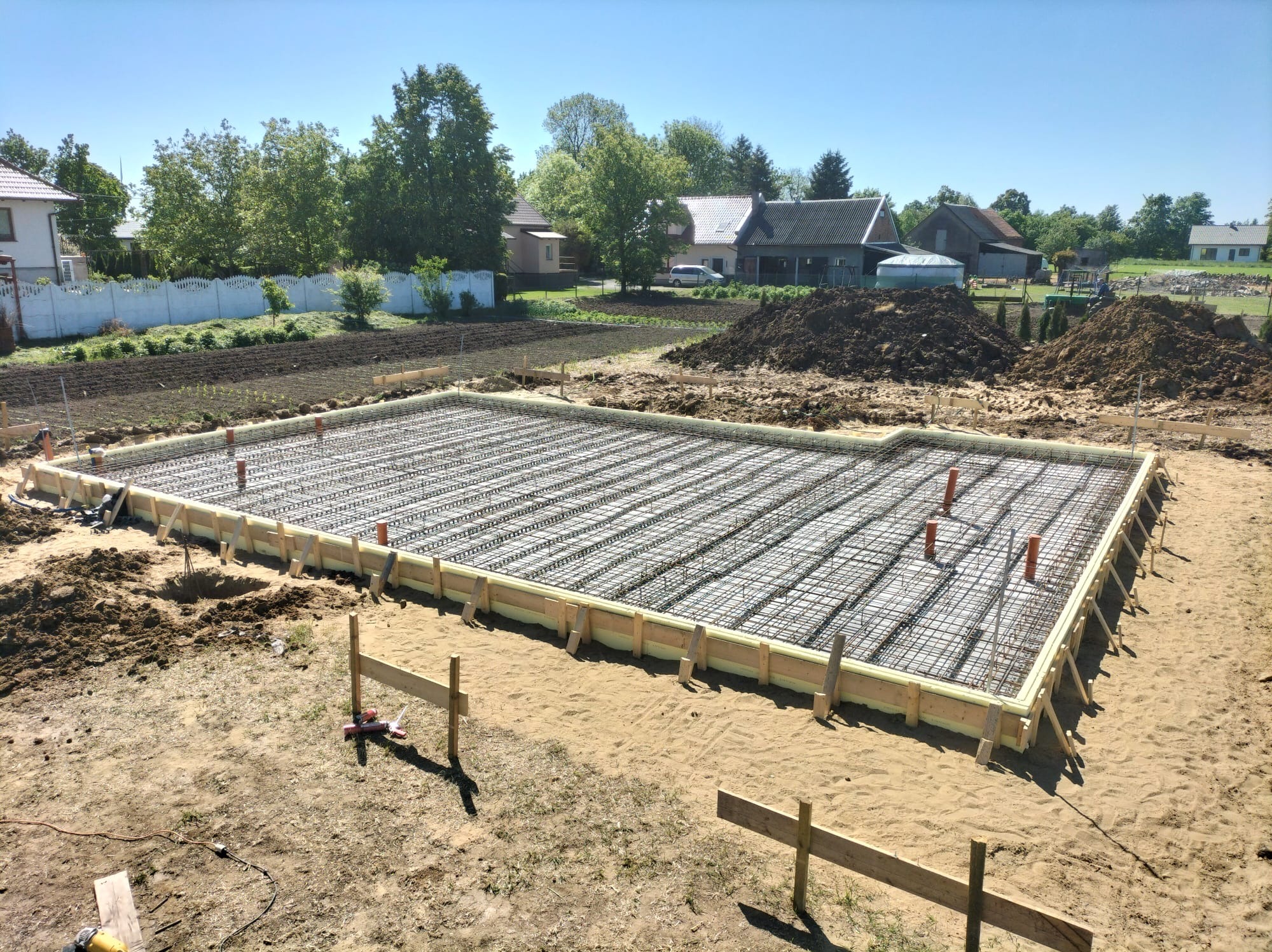Zbrojenie fundamentów pod dom jednorodzinny. Widoczne szalunki, izolacja i wystające rury. W tle zabudowa jednorodzinna i drzewa w słoneczny dzień.