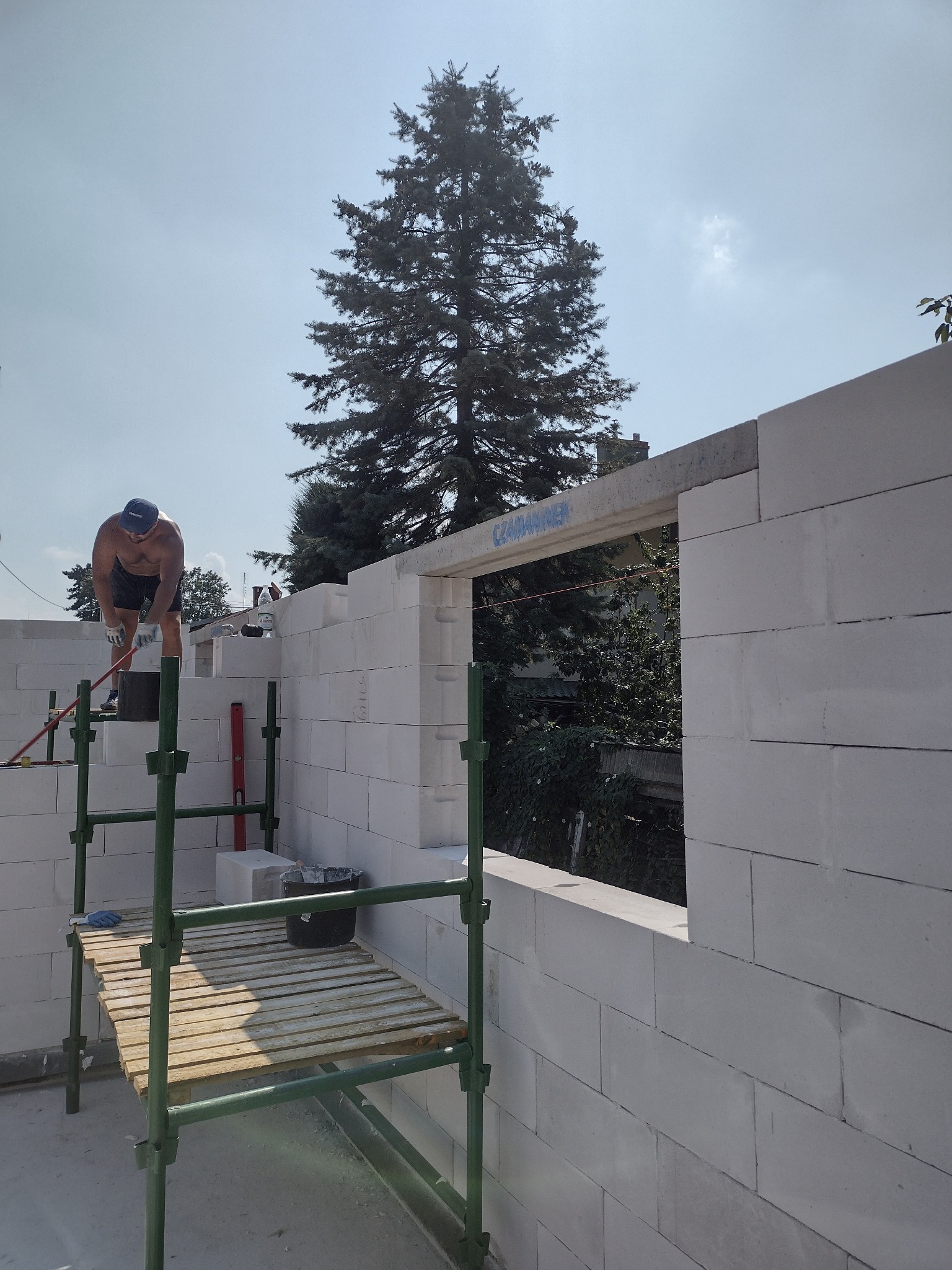 Mężczyzna w trakcie murowania ściany z białych bloczków na rusztowaniu, widoczny fragment okna z napisem, w tle drzewo iglaste.