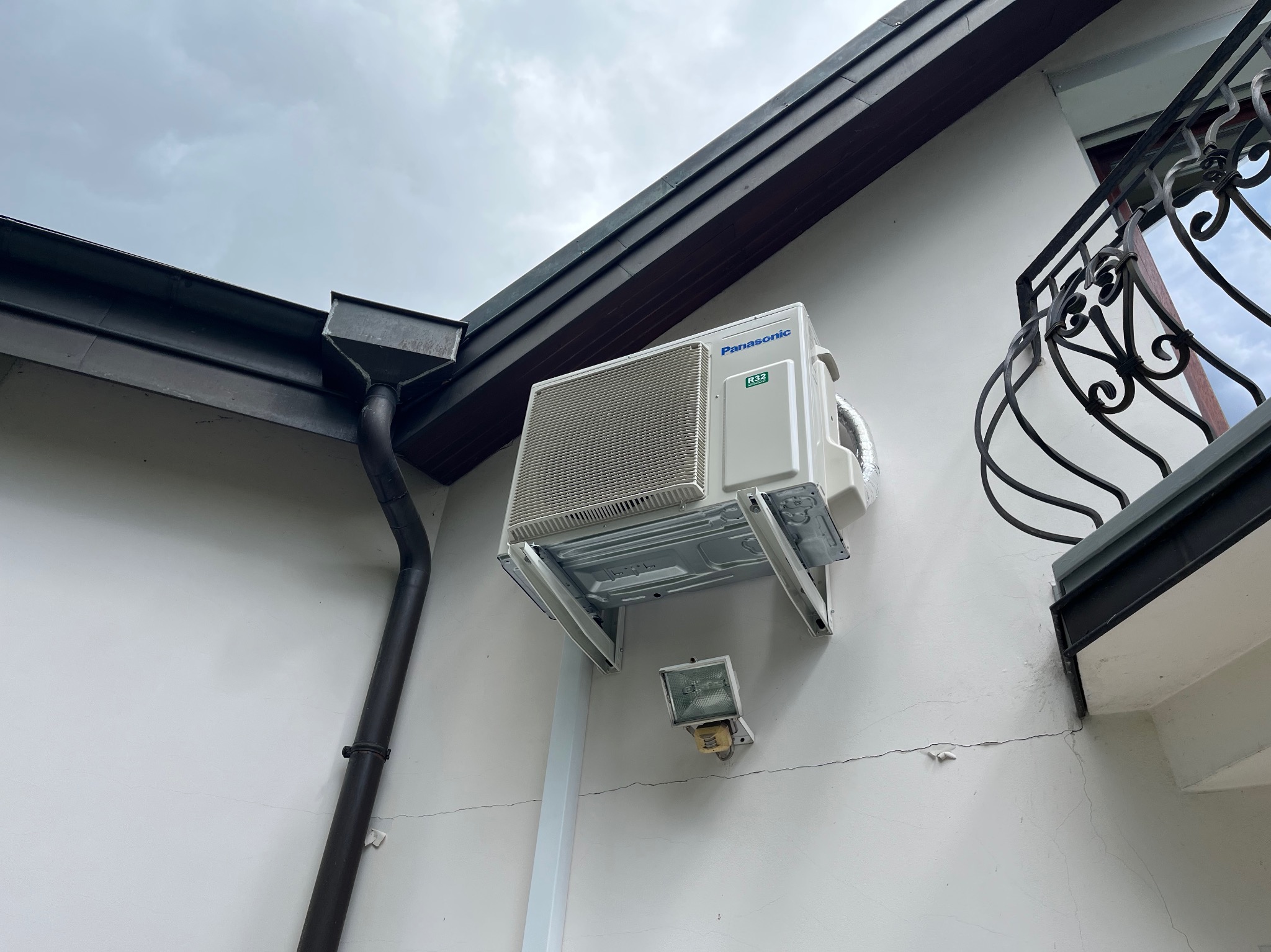 Zewnętrzna jednostka klimatyzacji Panasonic zamontowana na białej ścianie domu, widoczne rury odprowadzające wodę, metalowa rynna i fragment balkonu z kutą balustradą.