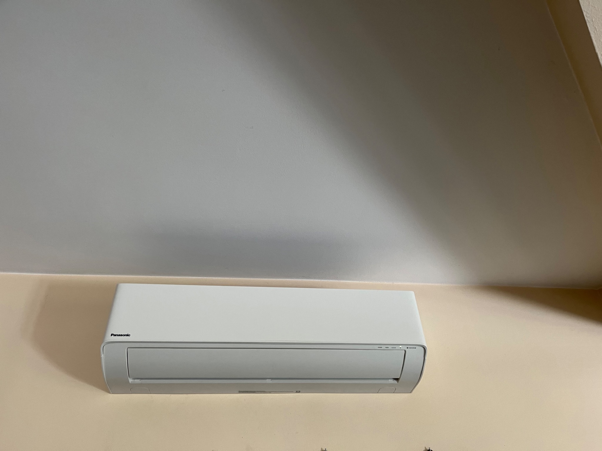 Biała jednostka wewnętrzna klimatyzacji Panasonic zamontowana na beżowej ścianie, widok od dołu, z widocznym fragmentem białego sufitu.