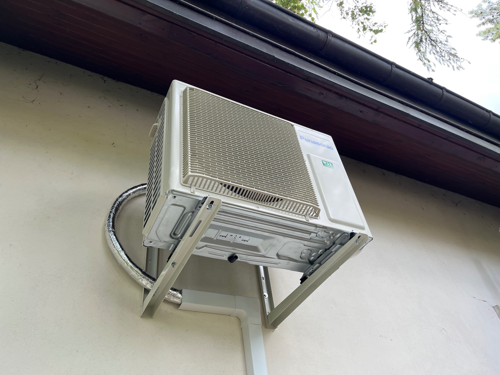Zewnętrzna jednostka klimatyzatora marki Panasonic R32 zamontowana na wspornikach na elewacji budynku, widok z dołu, z widocznymi przewodami w izolacji.