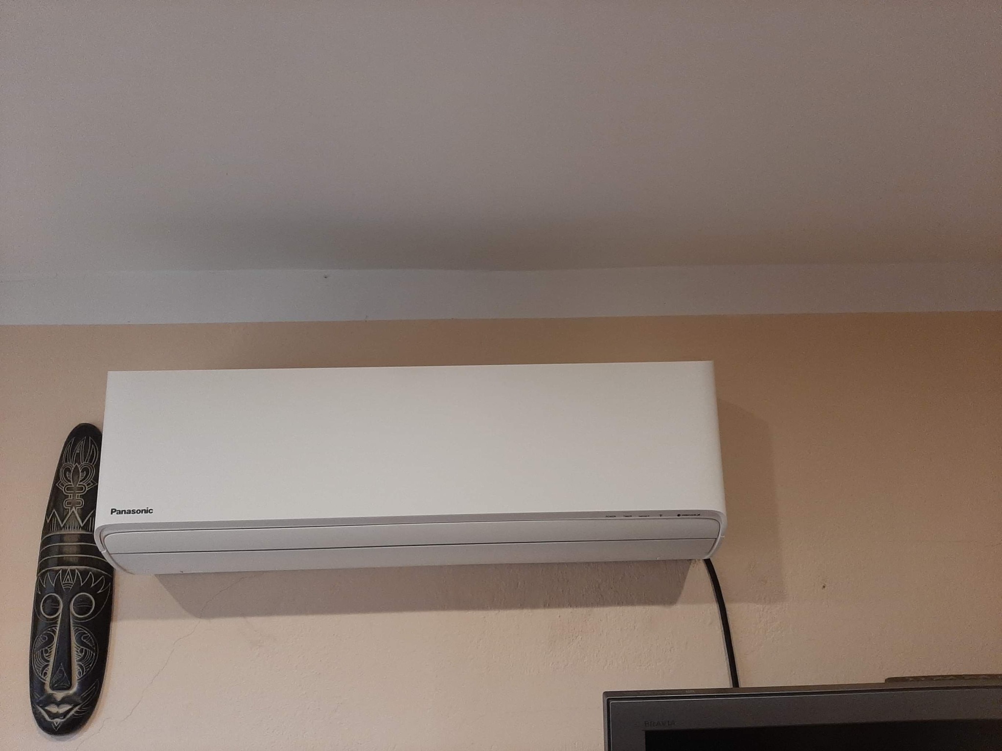 Biały klimatyzator Panasonic zamontowany na beżowej ścianie w pomieszczeniu, obok wisi afrykańska maska. Widoczny fragment telewizora.