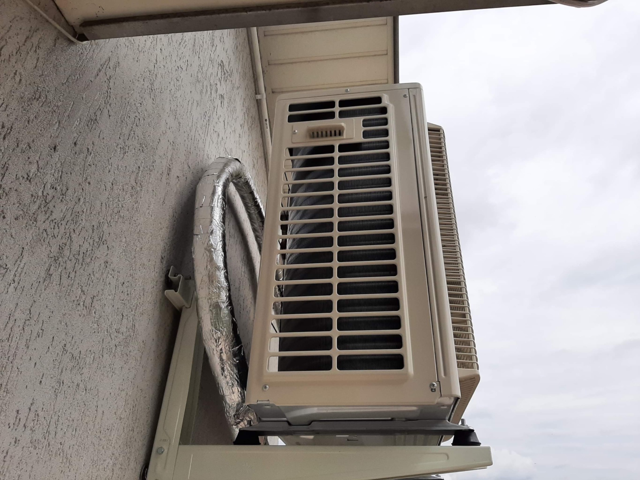 Zewnętrzna jednostka klimatyzatora zamontowana na ścianie budynku z widoczną rurą odprowadzającą skropliny owiniętą folią izolacyjną.