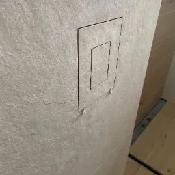 Ściana wyłożona beżowymi płytkami z ukrytym otworem rewizyjnym, widoczne małe plastikowe kliny do płytek i fragment odpływu liniowego.