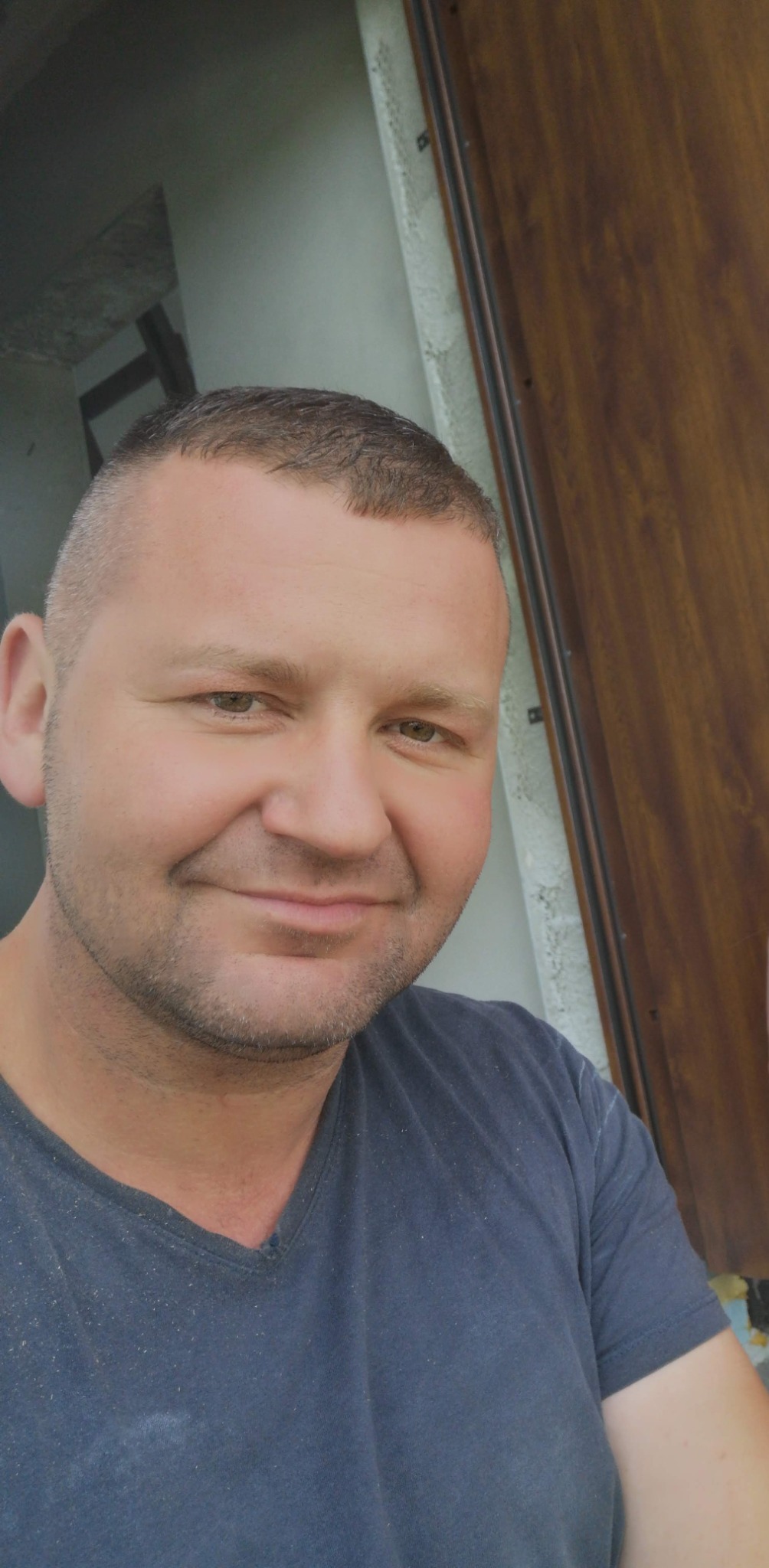 Portret uśmiechniętego mężczyzny w niebieskiej koszulce na tle częściowo widocznych drzwi i fragmentu ściany w trakcie prac wykończeniowych.