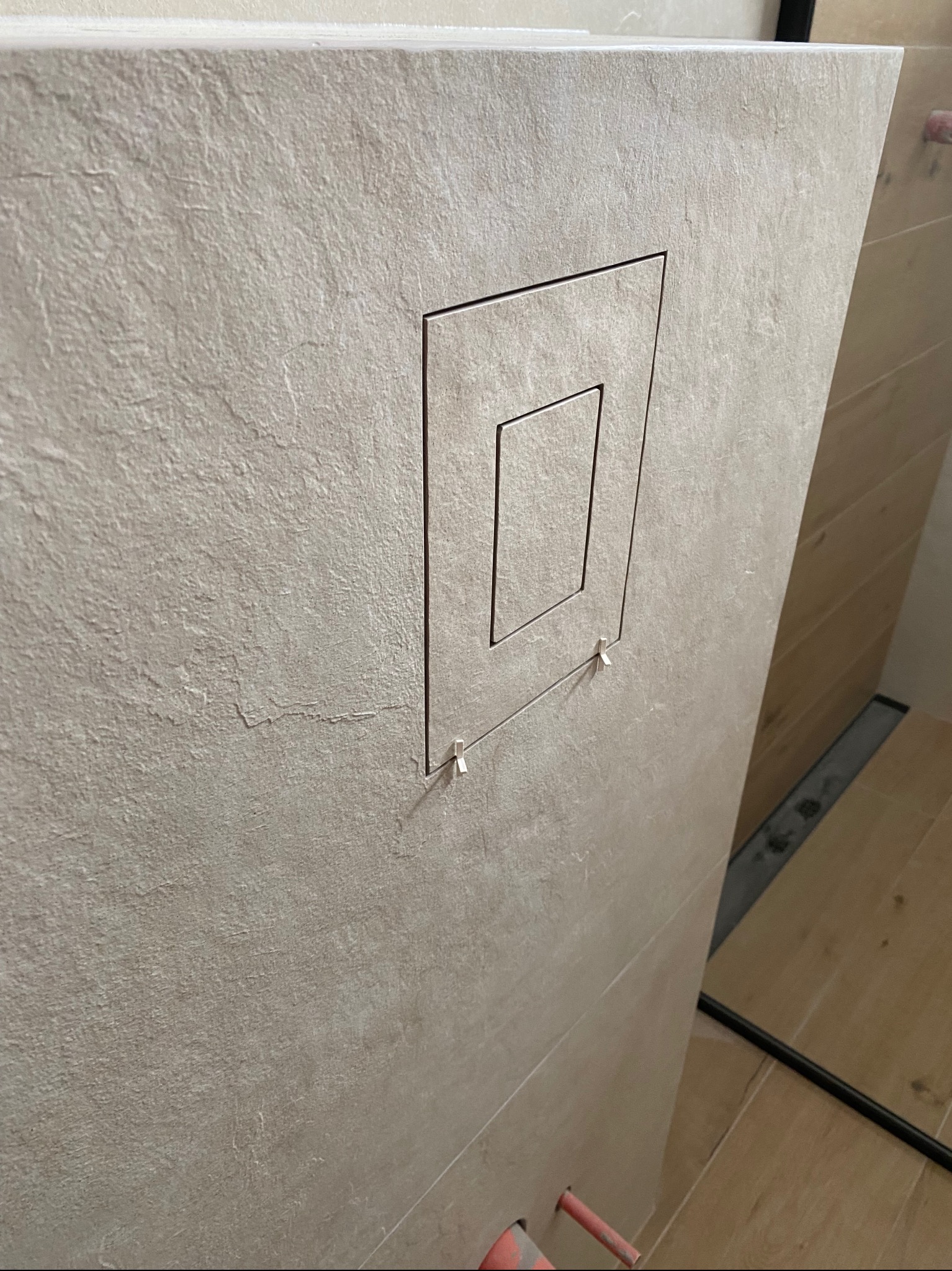 Ściana wyłożona beżowymi płytkami z ukrytym otworem rewizyjnym, widoczne małe plastikowe kliny do płytek i fragment odpływu liniowego.