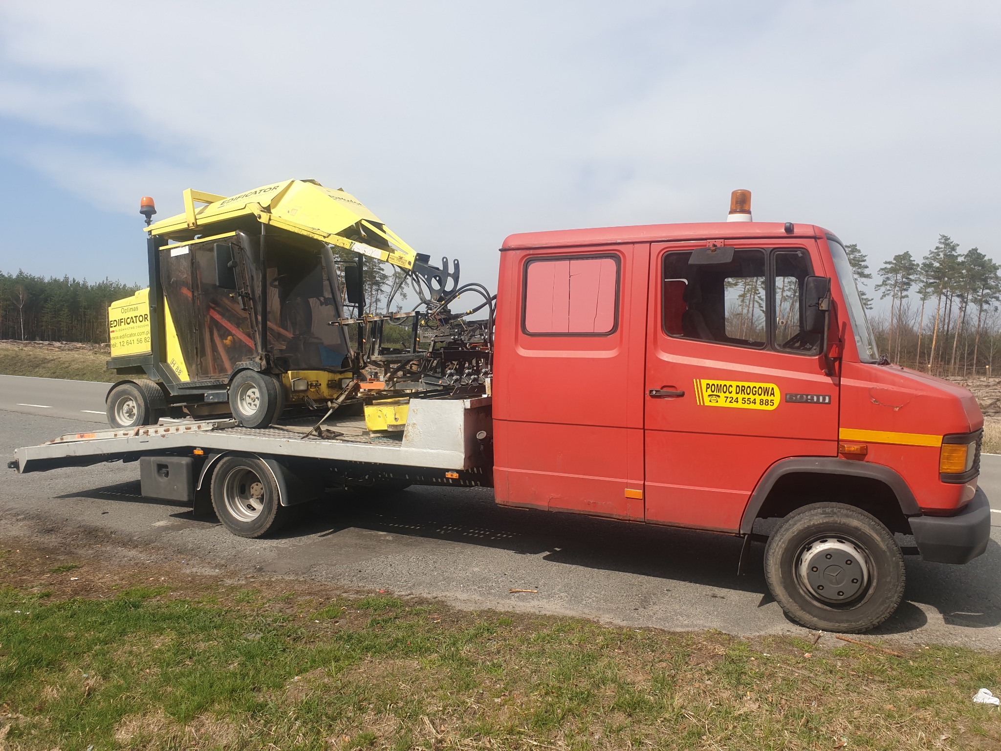 Czerwona laweta z żółtą frezarką do pni drzew na platformie, widoczne logo firmy Edificator na maszynie, tabliczka Pomoc Drogowa na kabinie lawety.