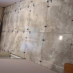 Robert Przybysławski - Szare płytki podłogowe w trakcie układania, z niebieskimi krzyżykami dystansowymi, widoczny fragment grzejnika i okna.