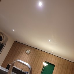 Robert Przybysławski - Wykończone wnętrze z drewnopodobną płytą ścienną, punktowym oświetleniem sufitowym i zegarem na ścianie. W tle widoczna drabina malarska i zasłonięte przejście.