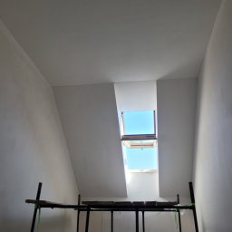 Robert Przybysławski - Pokój na poddaszu w trakcie prac wykończeniowych, widoczne rusztowanie, ściany i sufit po nałożeniu gładzi, otwarte okno dachowe wpuszcza jasne światło dzienne.