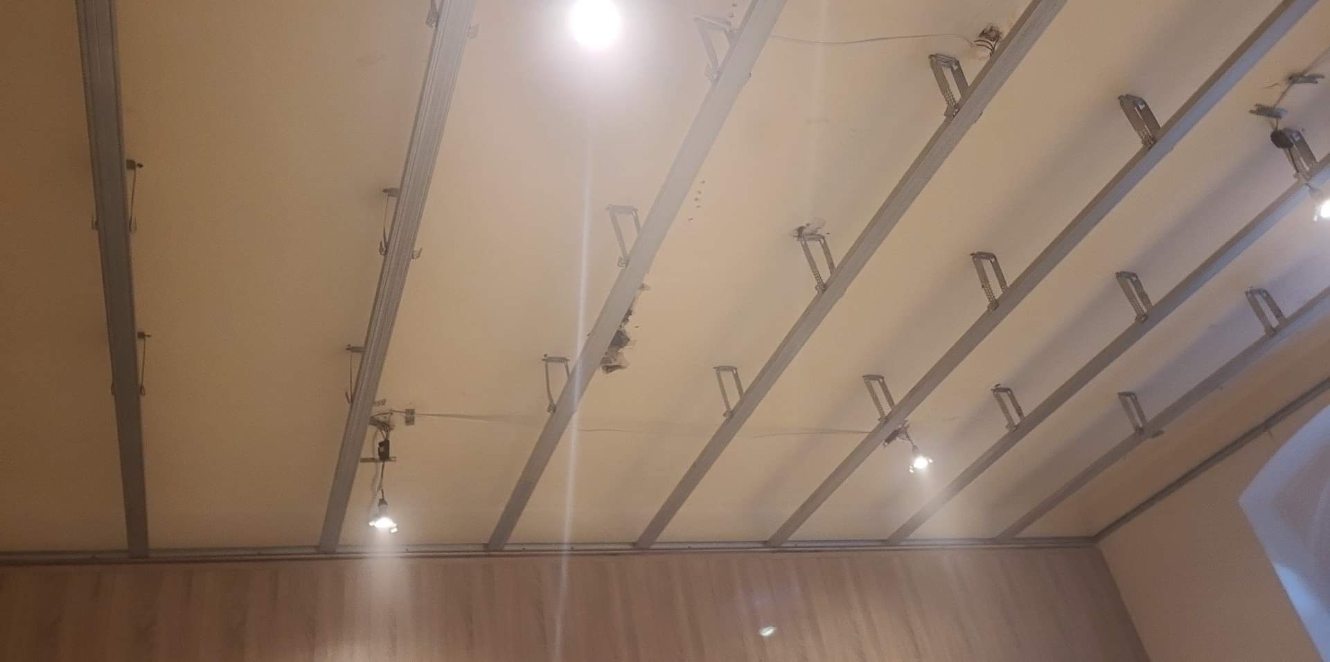 Metalowa konstrukcja podwieszanego sufitu z widocznymi elementami mocującymi i przewodami elektrycznymi, oświetlona przez żarówki, z fragmentem ściany w tle.