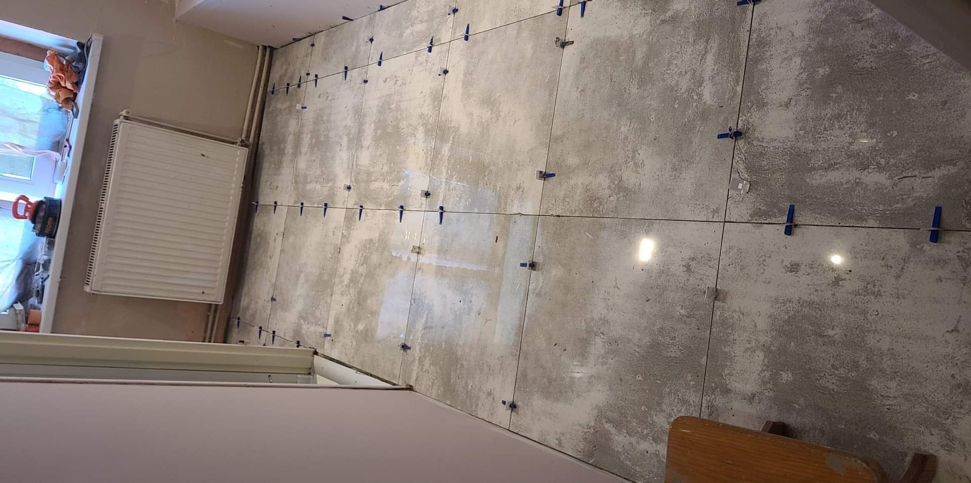 Szare płytki podłogowe w trakcie układania, z niebieskimi krzyżykami dystansowymi, widoczny fragment grzejnika i okna.