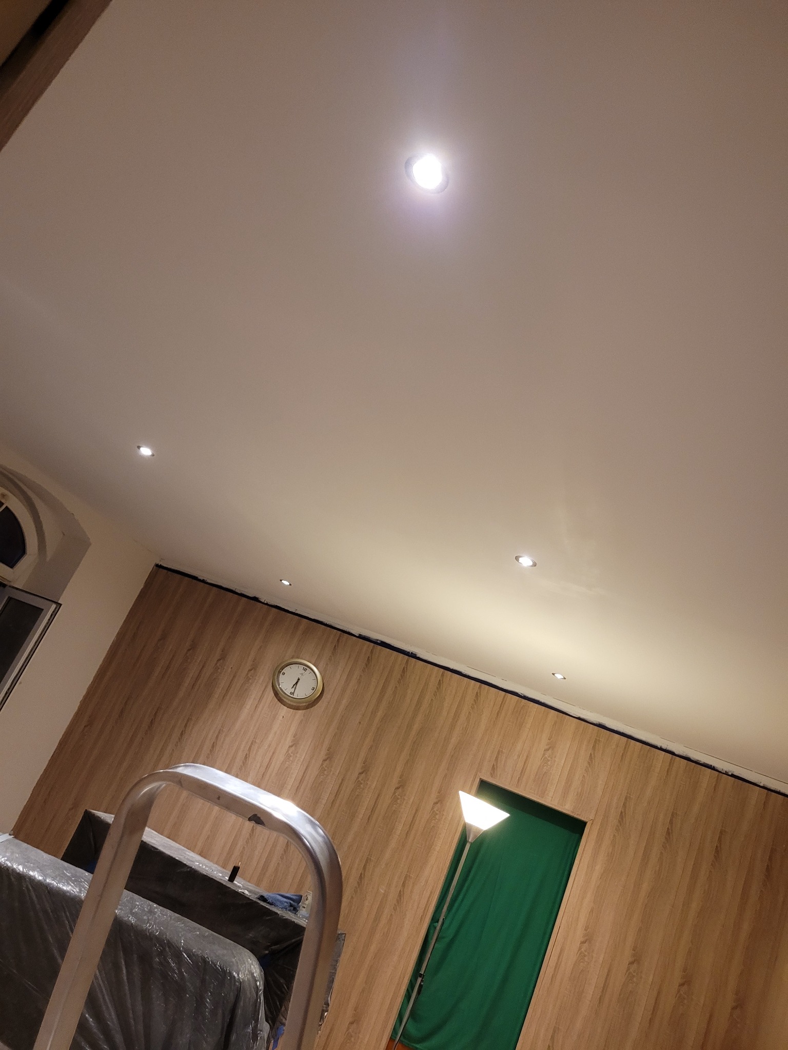 Wykończone wnętrze z drewnopodobną płytą ścienną, punktowym oświetleniem sufitowym i zegarem na ścianie. W tle widoczna drabina malarska i zasłonięte przejście.