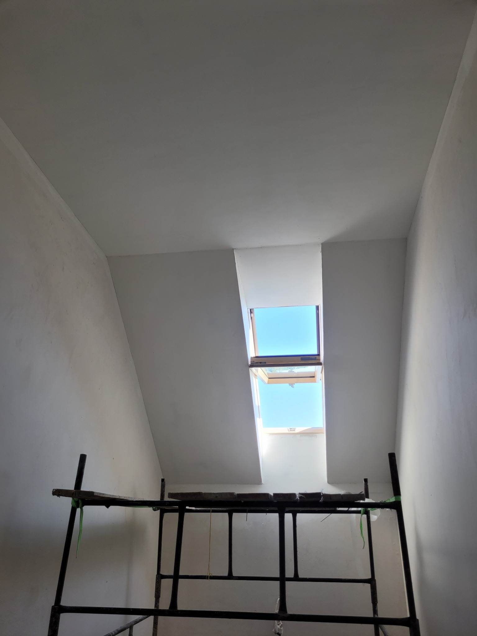 Pokój na poddaszu w trakcie prac wykończeniowych, widoczne rusztowanie, ściany i sufit po nałożeniu gładzi, otwarte okno dachowe wpuszcza jasne światło dzienne.