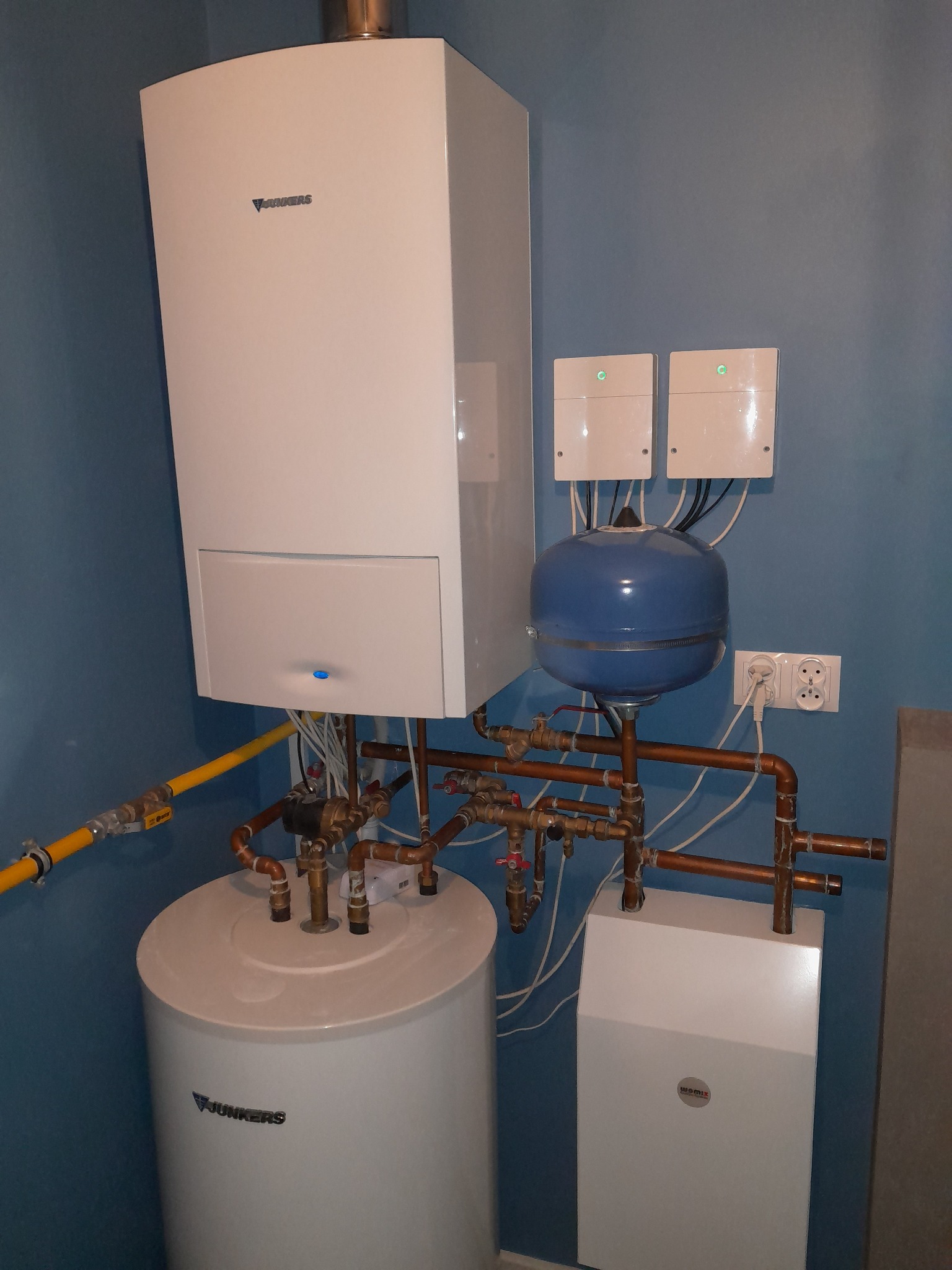 Kompaktowa kotłownia: biały piec gazowy Junkers, zasobnik ciepłej wody, niebieski zbiornik wyrównawczy i miedziane rury na tle błękitnej ściany.