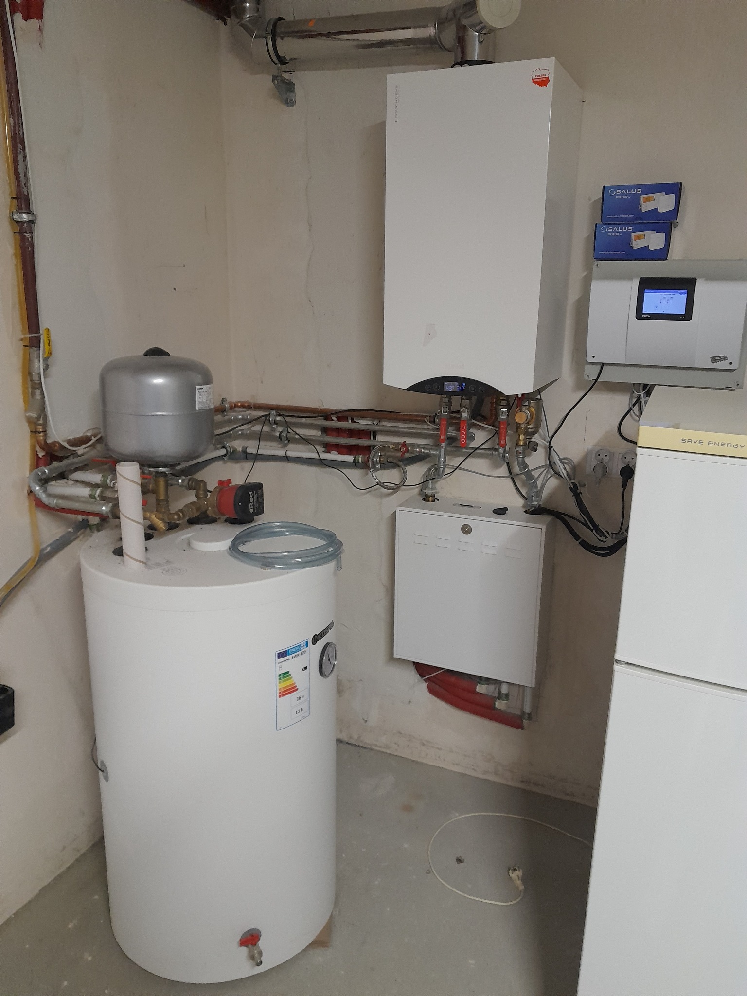 Nowoczesna instalacja grzewcza w kotłowni: biały, wiszący piec gazowy z podłączonymi rurami, zbiornik wyrównawczy, zasobnik ciepłej wody z etykietą energetyczną, sterownik Salus i dodatkowy panel...