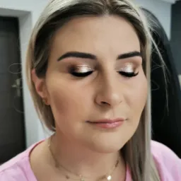 Portret kobiety z zamkniętymi oczami prezentującej złoty, błyszczący cień do powiek, delikatny róż na policzkach i błyszczyk na ustach. Widoczna dwuwarstwowa złota kolia.