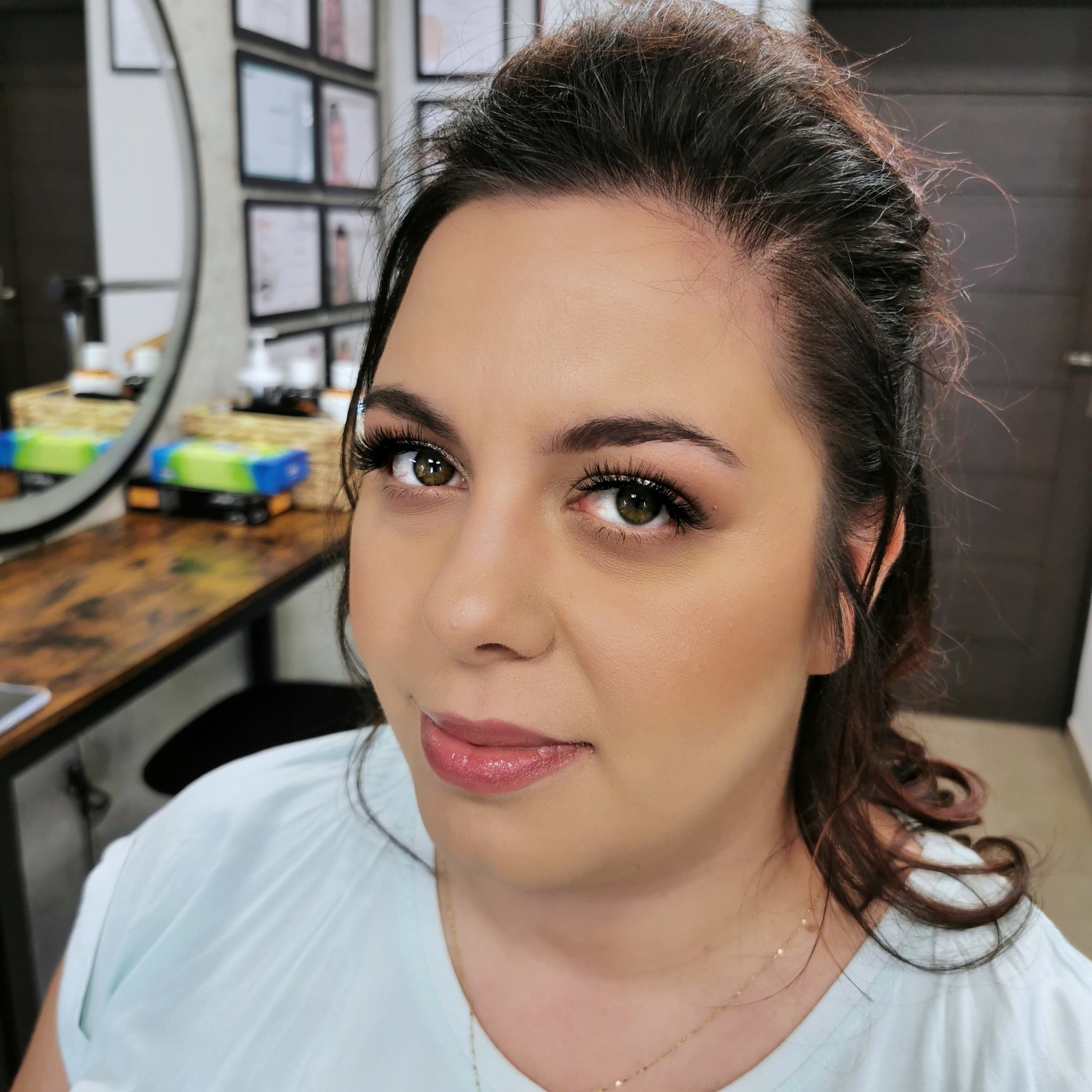 Portret kobiety z delikatnym, rozświetlonym makijażem, podkreślonymi rzęsami i błyszczącymi ustami, na tle lustra i certyfikatów w salonie.