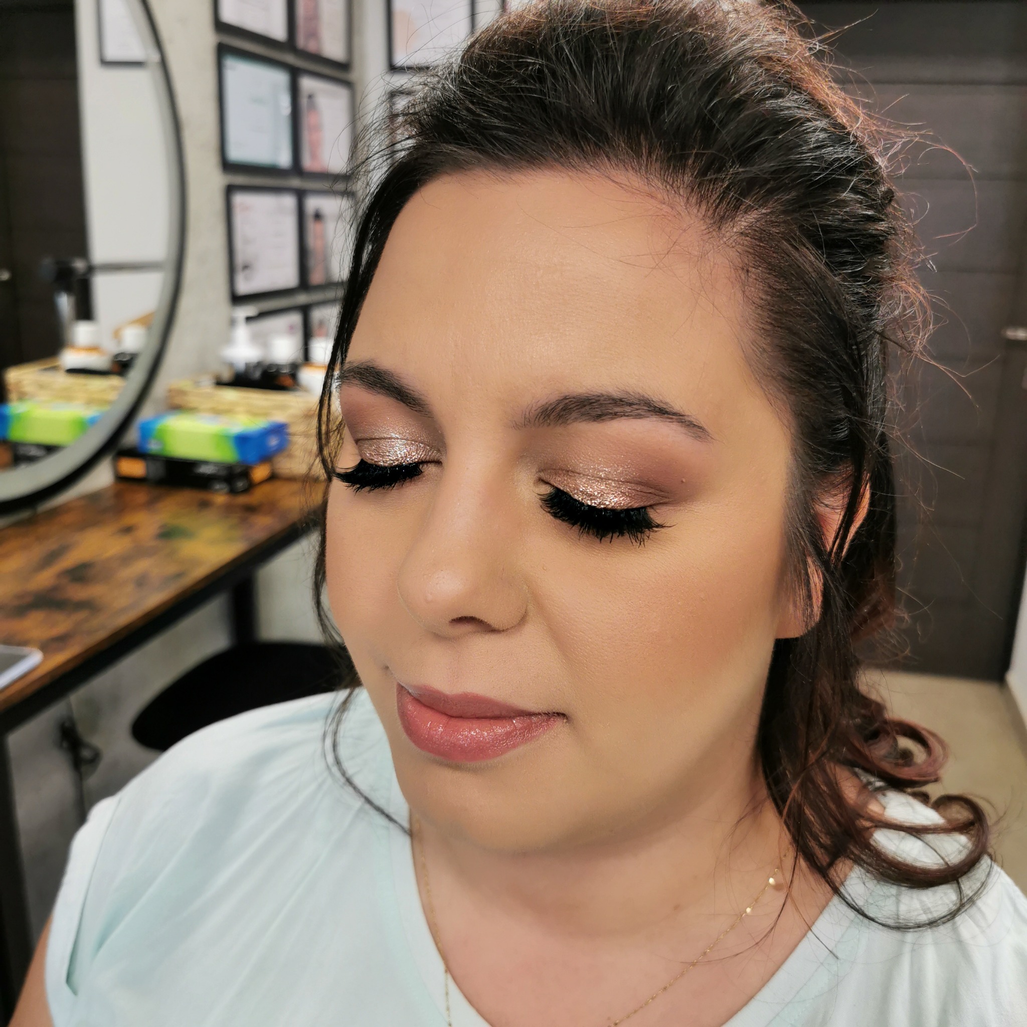 Portret kobiety z zamkniętymi oczami, prezentujący delikatny, błyszczący cień do powiek w odcieniach złota i brązu oraz wydłużone rzęsy. Usta podkreślone błyszczykiem w kolorze nude. W tle widoczne...