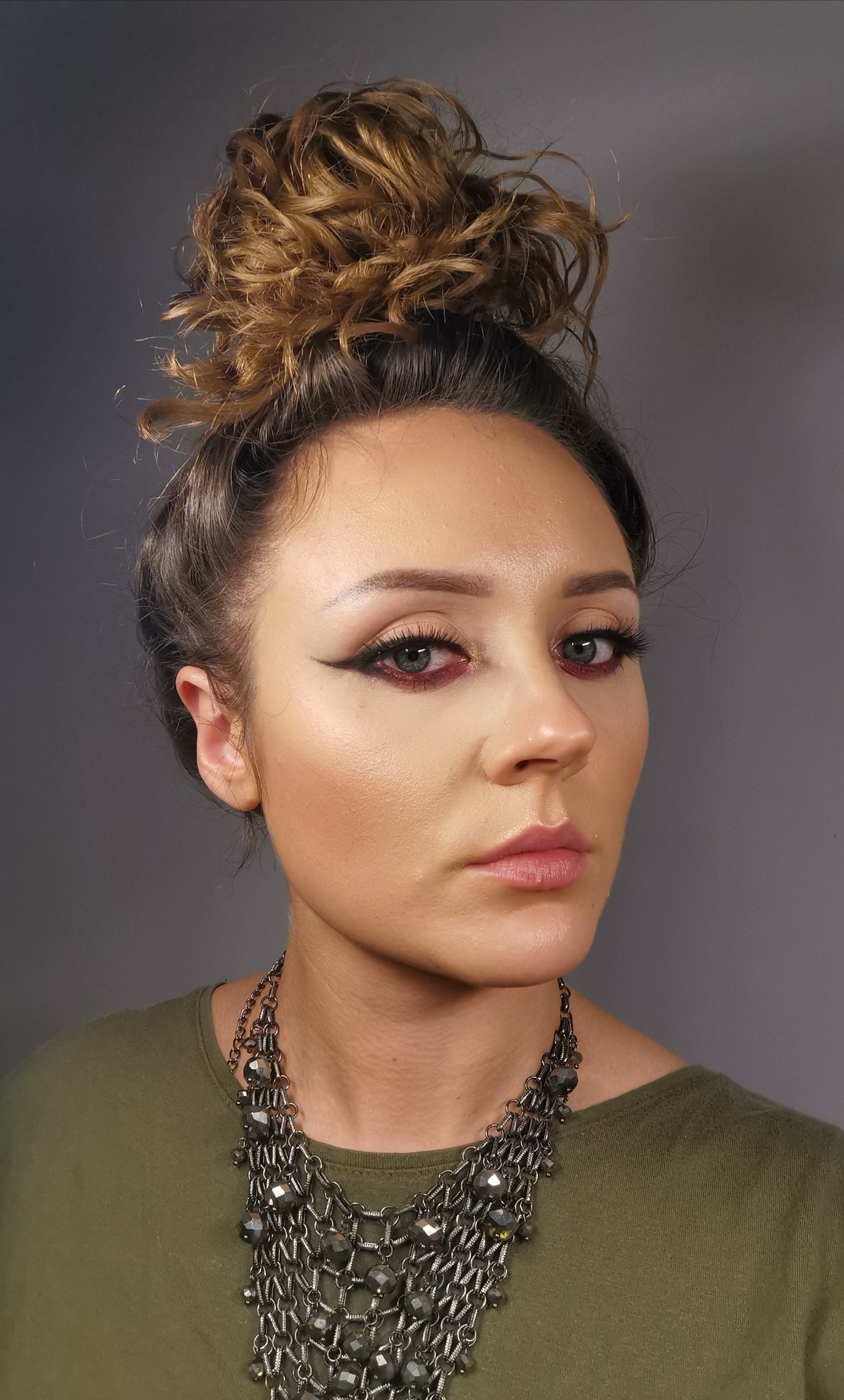Portret kobiety z intensywnym makijażem oczu w odcieniach bordo i czerni, włosy upięte w wysoki kok, ubrana w oliwkową koszulkę i masywny, metalowy naszyjnik.