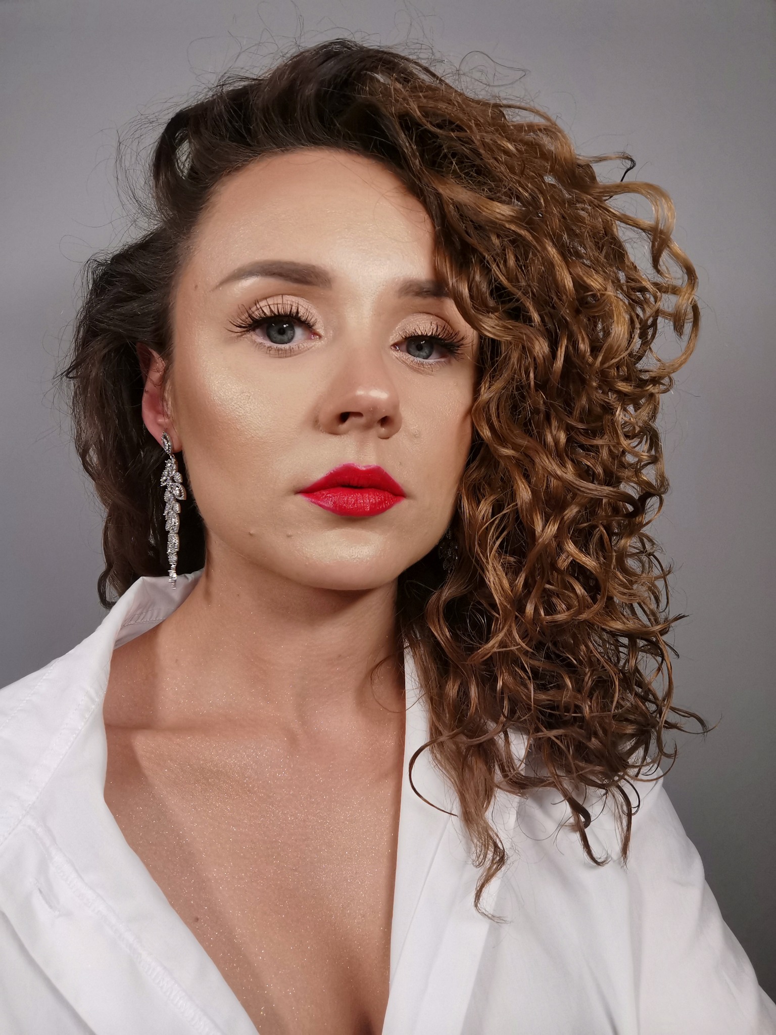 Portret kobiety z wyrazistym makijażem ust w kolorze czerwieni, podkreślonymi rzęsami i długimi kolczykami w kształcie gałązek na tle szarego studia.