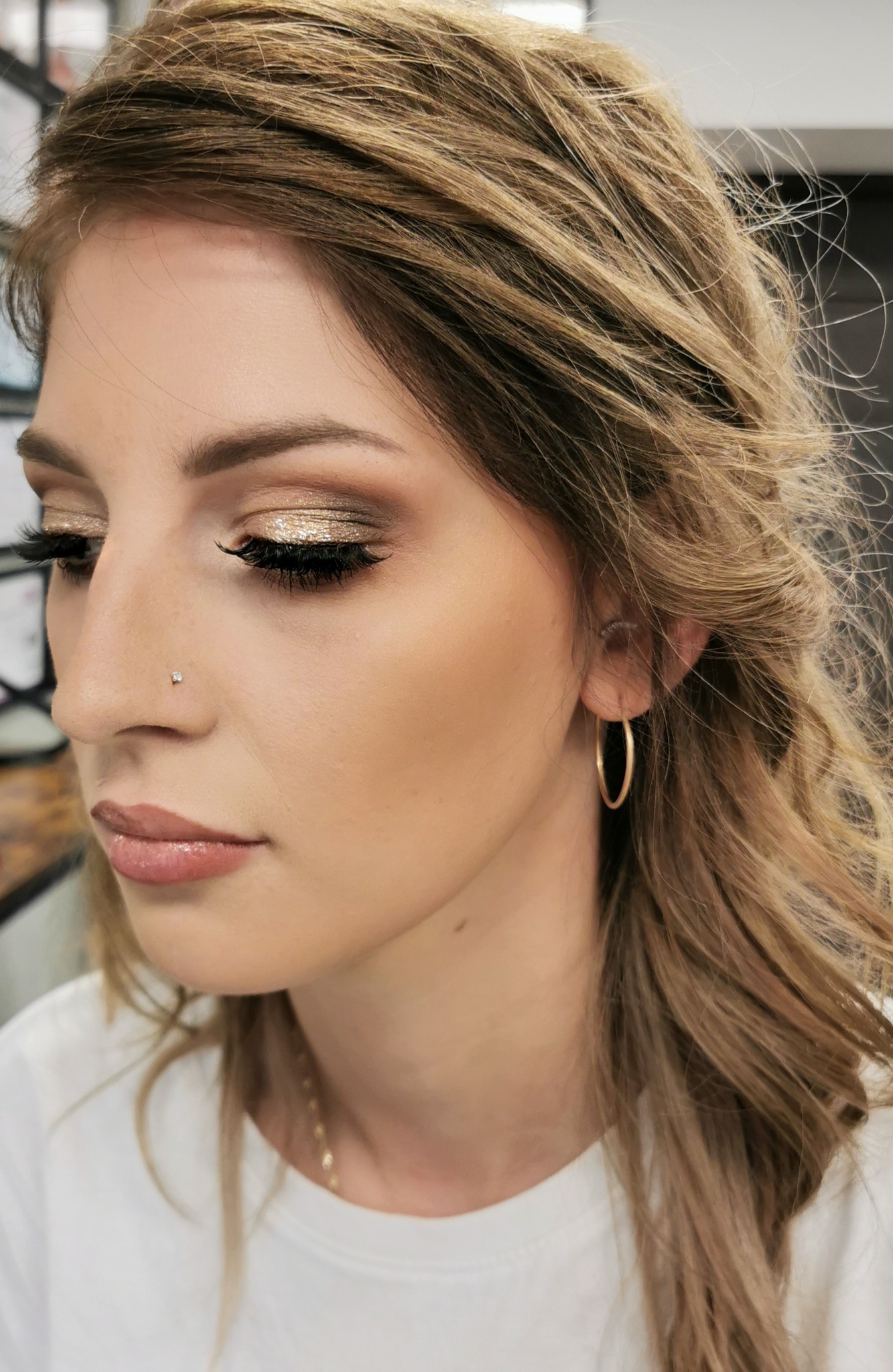 Profil kobiety z blond włosami i złotym, brokatowym cieniem na powiekach oraz delikatnym makijażem ust i skóry, z kolczykiem w nosie i złotym kolczykiem w uchu.