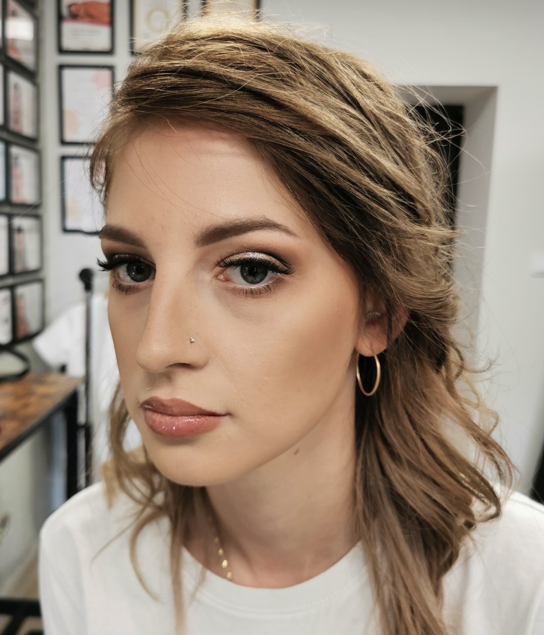 Portret kobiety z delikatnym, błyszczącym makijażem oczu w odcieniach złota i brązu, podkreślonymi rzęsami i błyszczącymi ustami, z kolczykiem w nosie i złotymi kolczykami kołami.