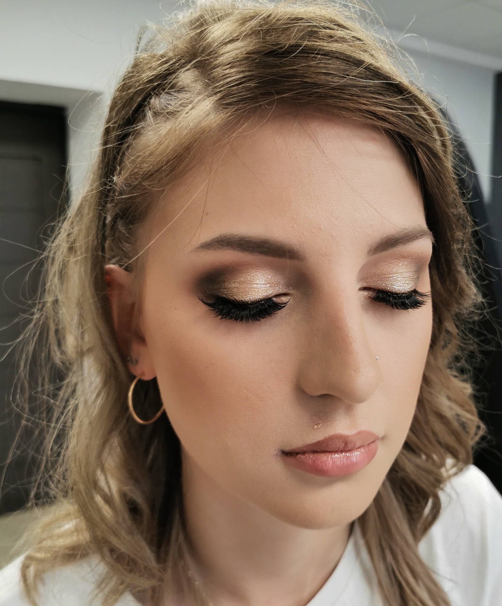 Portret kobiety z zamkniętymi oczami prezentujący efektowny, złoty makijaż oczu z długimi, sztucznymi rzęsami, wykonany w salonie w Wałbrzychu. Widoczne delikatne kolczyki koła i subtelny błyszczyk...