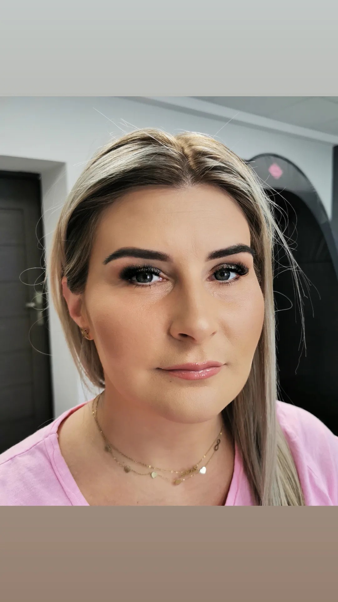 Portret kobiety z profesjonalnym makijażem oczu i ust w odcieniach nude, z delikatną biżuterią, na rozmytym tle salonu.