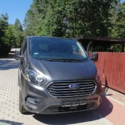 Szary Ford Transit Custom z otwartymi drzwiami kierowcy, zaparkowany na kostce brukowej przed czerwonym płotem i zielonymi drzewami w słoneczny dzień.