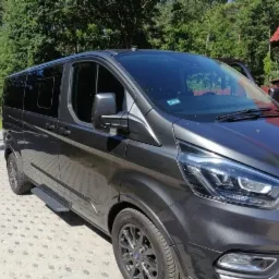 Szary, luksusowy van z przyciemnianymi szybami, zaparkowany na betonowej kostce brukowej, z otwartymi drzwiami po stronie kierowcy, na tle zieleni i czerwonego płotu.