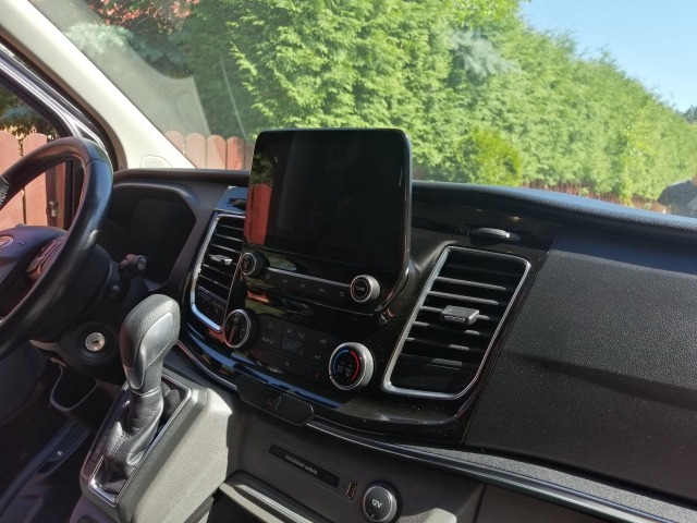 Wnętrze samochodu dostawczego Ford Transit z automatyczną skrzynią biegów, widoczny kokpit z ekranem dotykowym i kratki wentylacyjne na tle zieleni.