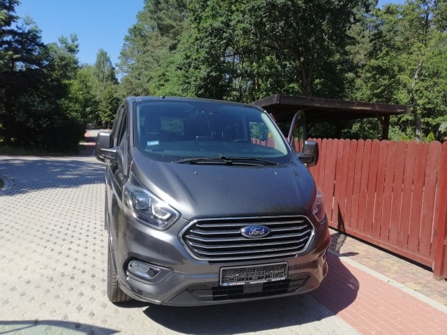 Szary Ford Transit Custom z otwartymi drzwiami kierowcy, zaparkowany na kostce brukowej przed czerwonym płotem i zielonymi drzewami w słoneczny dzień.