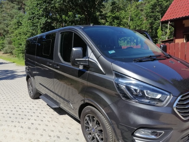 Szary, luksusowy van z przyciemnianymi szybami, zaparkowany na betonowej kostce brukowej, z otwartymi drzwiami po stronie kierowcy, na tle zieleni i czerwonego płotu.