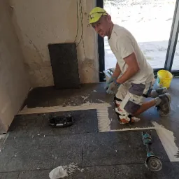 Glazurnik w trakcie układania ciemnych płytek podłogowych z krzyżykami dystansowymi, widoczna szlifierka kątowa i narzędzia glazurnicze.