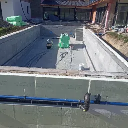 Budowa basenu zewnętrznego w ogrodzie: widok na częściowo wykończoną konstrukcję z izolacją termiczną, widoczne rury instalacyjne, w tle dom z dużymi oknami i materiały budowlane.