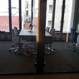 Nowoczesne biuro za szklaną ścianą z dekoracyjnymi wzorami, widoczne stoły konferencyjne, krzesła biurowe na kółkach i wykładzina w ciemnym kolorze.