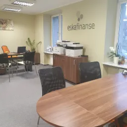 Biuro firmy z logo 'eskafinanse' na ścianie, widoczne biurka, krzesła, drukarki i roślina doniczkowa na parapecie okna.