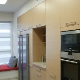 Jasne, nowoczesne meble kuchenne z drewnopodobnym fornirem, zabudowana lodówka, piekarnik i mikrofalówka, szary marmurkowy blat i podłoga, okno z roletą i siedziskiem.