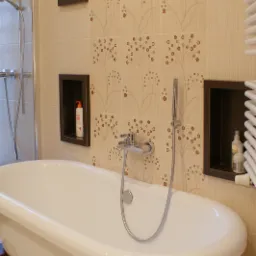 Wolnostojąca wanna na ciemnych, drewnianych nóżkach, bateria wannowa z prysznicem, beżowe płytki z motywem roślinnym i wnęki w ciemnych obramowaniach z kosmetykami.