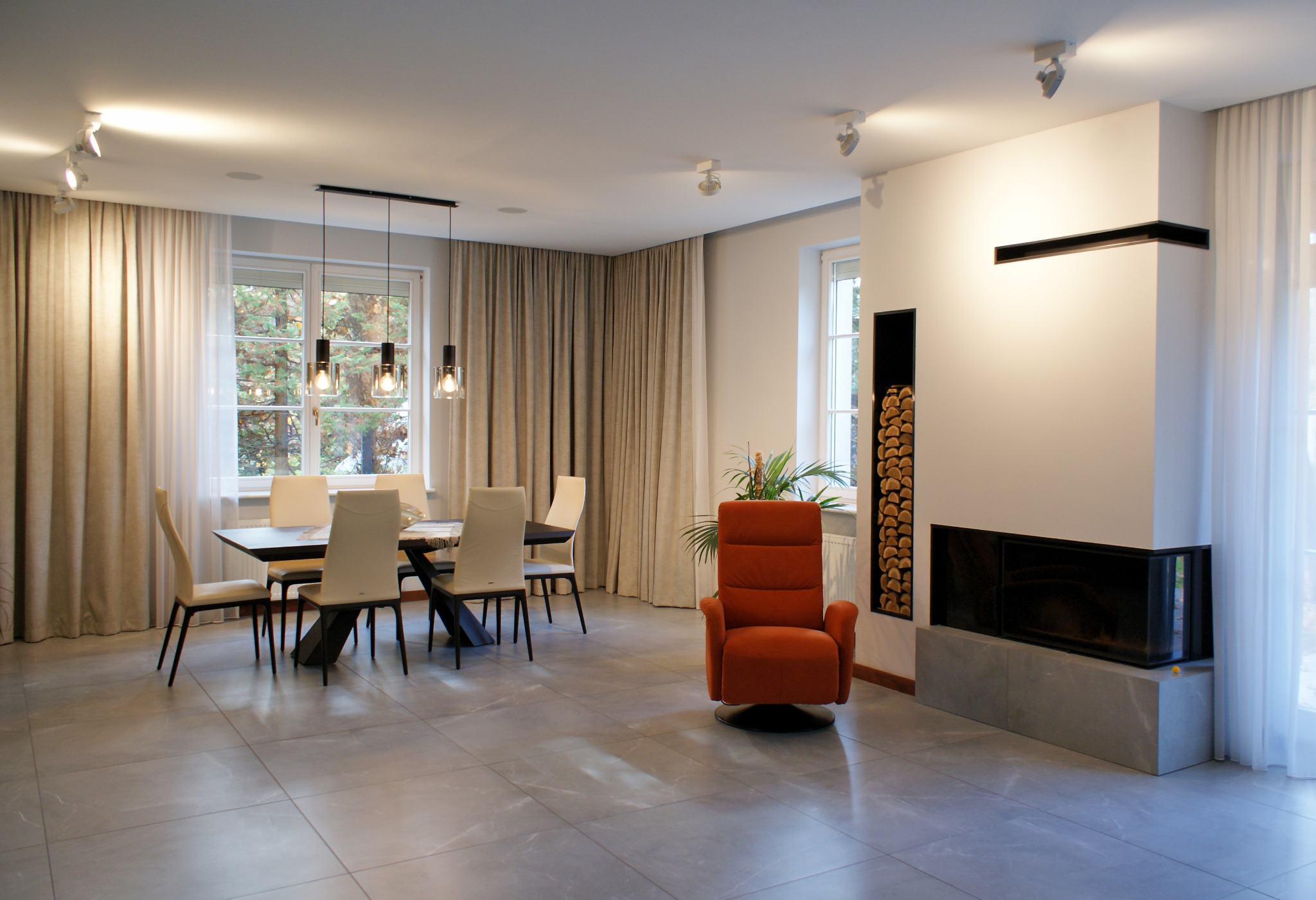 Jasne wnętrze jadalni z szarymi płytkami podłogowymi, stołem z krzesłami, pomarańczowym fotelem obrotowym i nowoczesnym kominkiem z drewnem w niszy.