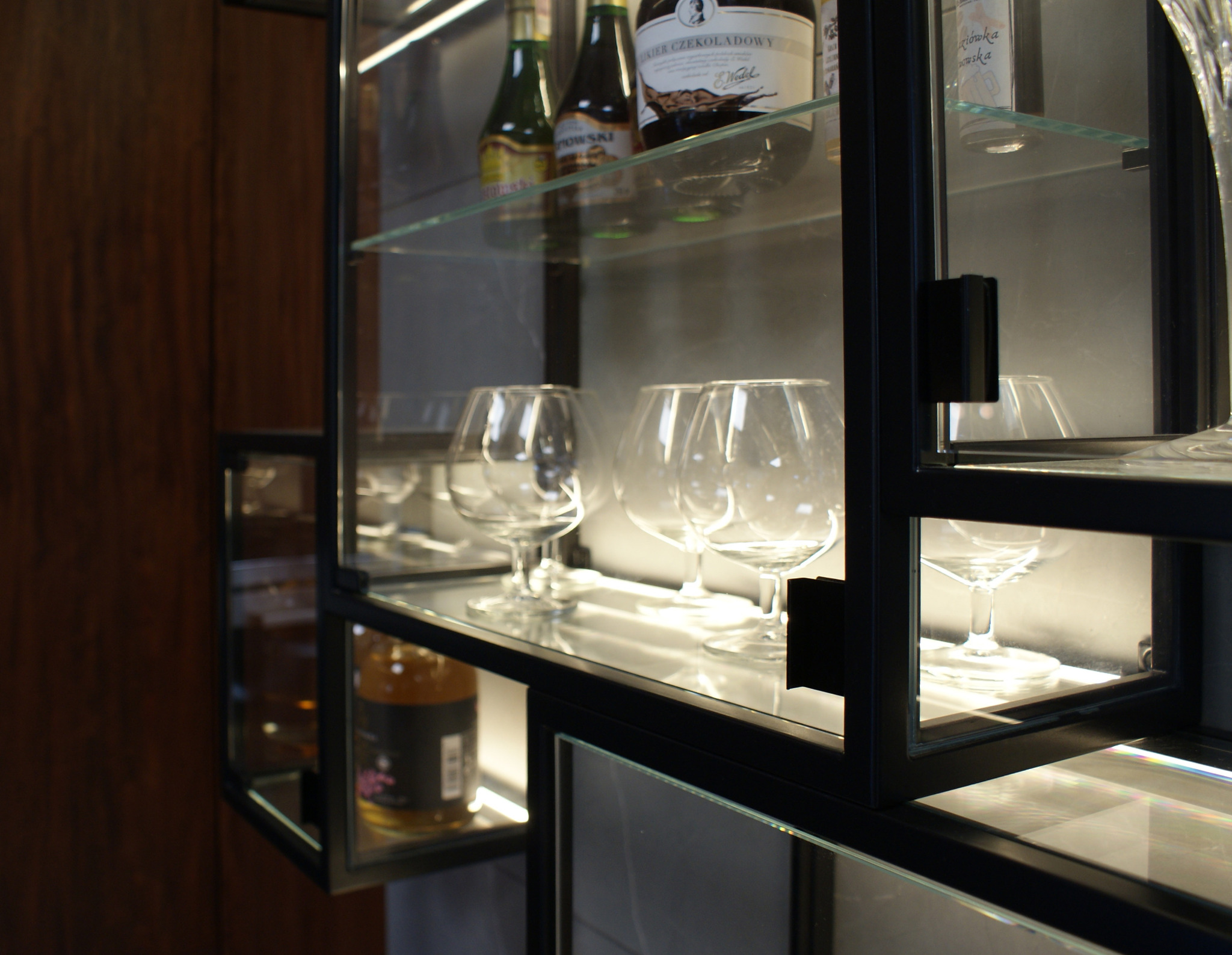 Szklana witryna z czarnymi ramami, podświetlona, prezentująca kolekcję kieliszków do wina i butelek alkoholu.