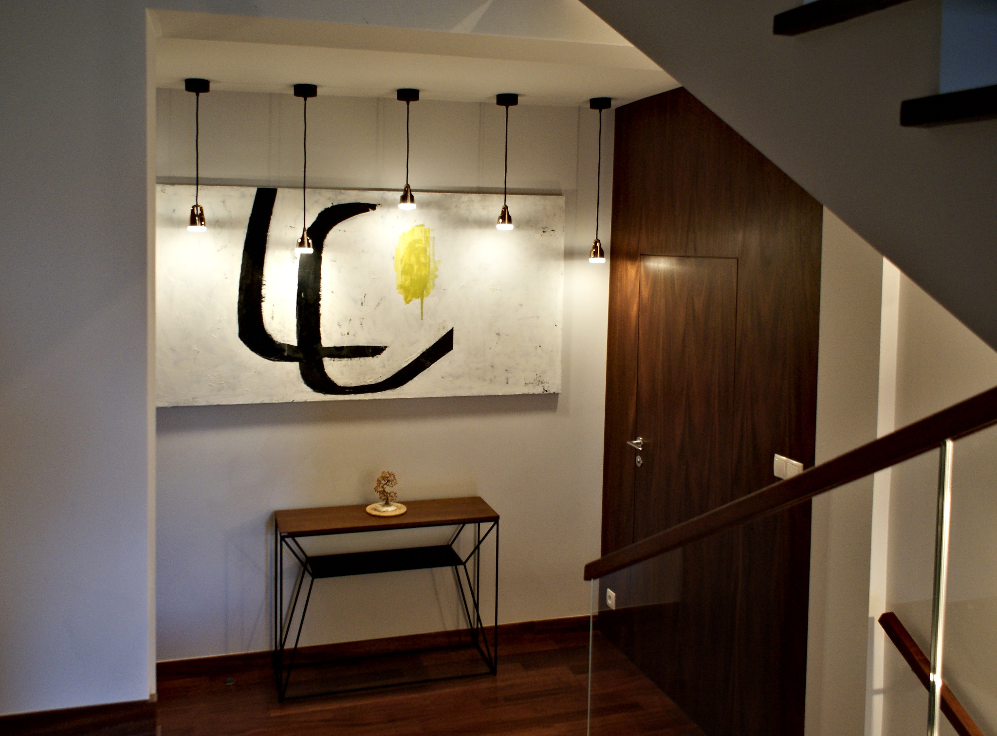 Nowoczesny przedpokój z drewnianą podłogą, konsolą z dekoracją, abstrakcyjnym obrazem z czarnymi i żółtymi akcentami, oświetlony wiszącymi lampami i z drewnianymi drzwiami w tle.