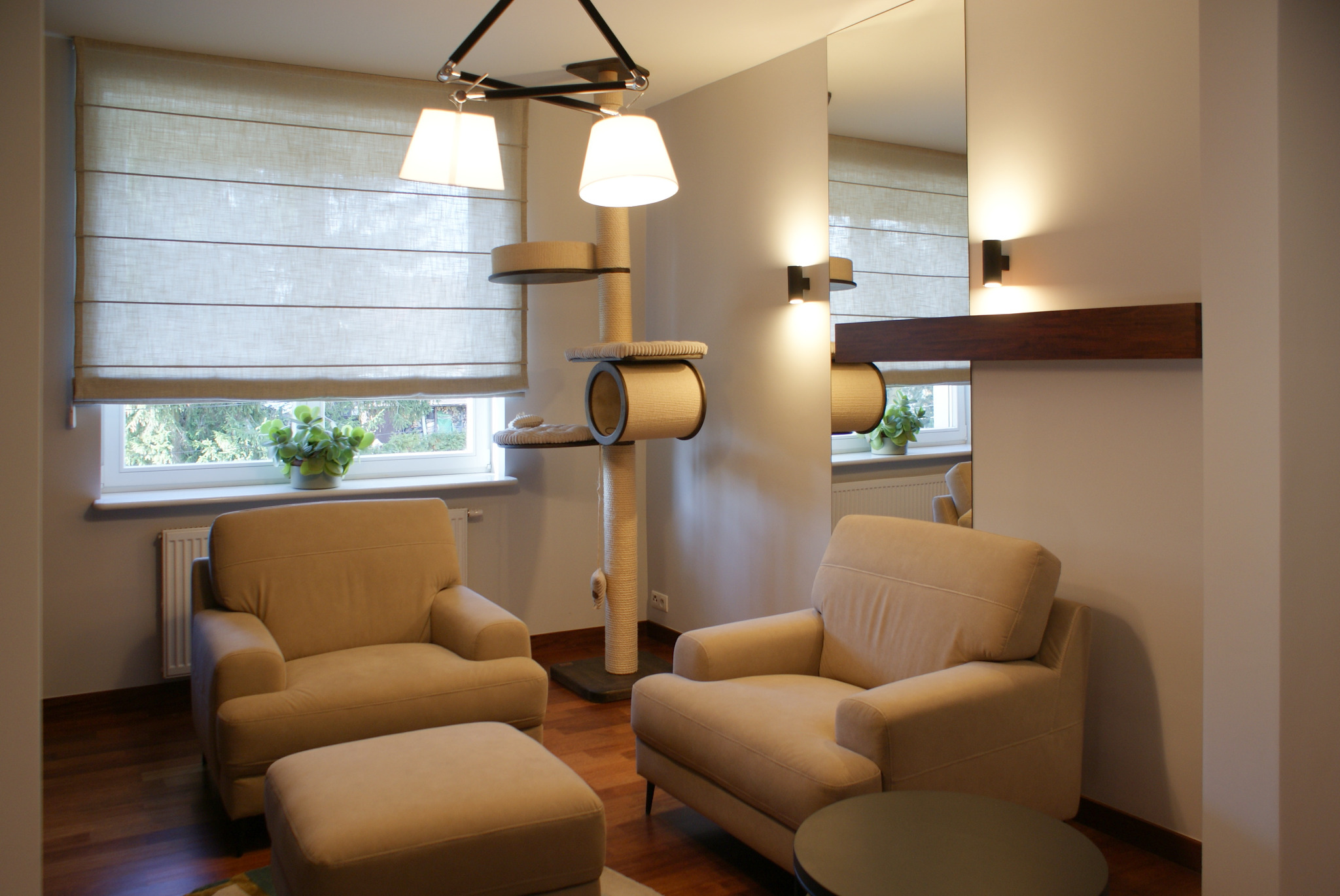 Jasny salon z dwoma fotelami, pufą, drapakiem dla kota, roletami rzymskimi i rośliną doniczkową na parapecie, odbicie w lustrze